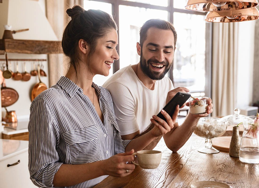 Read Our Reviews - Young Couple Smiles as They Use a Smartphone in Their Bright Kitchen, Enjoying Coffee