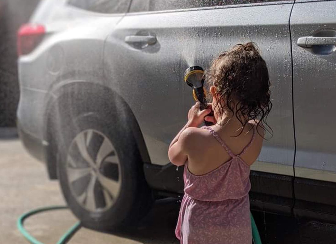 Personal Insurance - Little Girl is Using a Water Hose to Spray a Car With Water on a Sunny Day